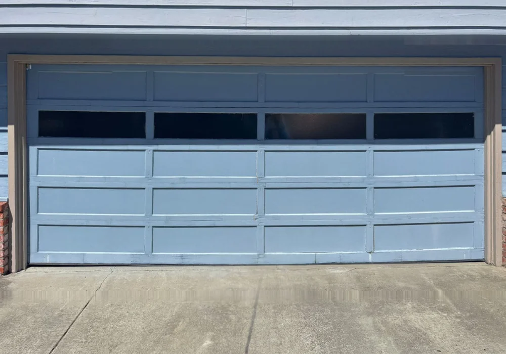 Worn pale blue sectional garage door with a row of windows, before replacement